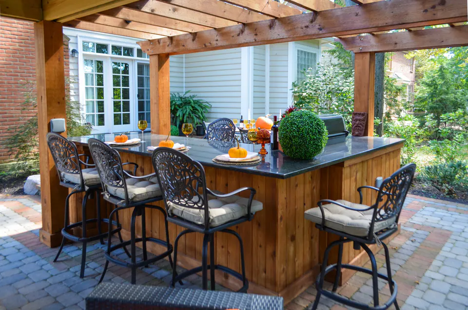 Outdoor dining table and wood bar beside house with pergola framing, paver patio, and fall table setting.